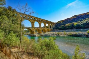 The ancient Roman aqueduct Pont du Gard, spanning a tranquil river surrounded by lush greenery and hills, under a bright blue sky.