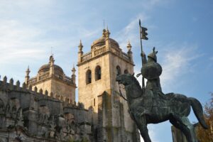 A statue of a medieval knight on horseback in front of the historic Porto Cathedral, featuring its distinctive towers and battlements, under a clear blue sky.