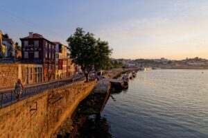 A scenic view of the Douro River waterfront in Porto at sunset, with colorful buildings, a tree-lined promenade, and boats docked along the river.