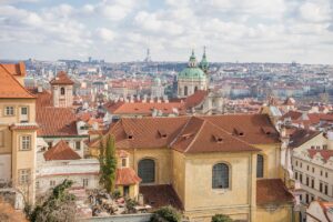 Panoramic view of Prague's historic cityscape with St. Nicholas Church and red-tiled rooftops under a partly cloudy sky.