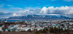 Panoramic view of Reykjavik, Iceland, with colorful buildings in the foreground and snow-capped mountains under a blue sky with clouds in the background.