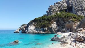 Crystal clear turquoise waters and rugged rocky cliffs at a secluded beach in Sardinia, Italy, with a few people enjoying the serene landscape.